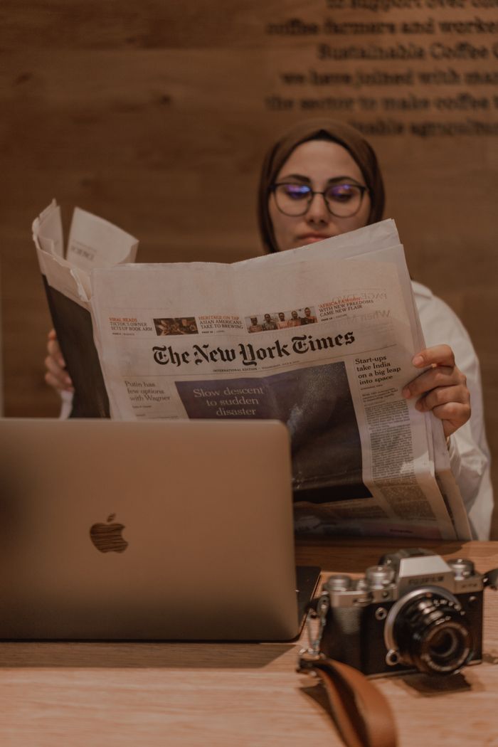 A Muslim woman in hijab reads a newspaper at a coffee shop, alongside a laptop and camera.