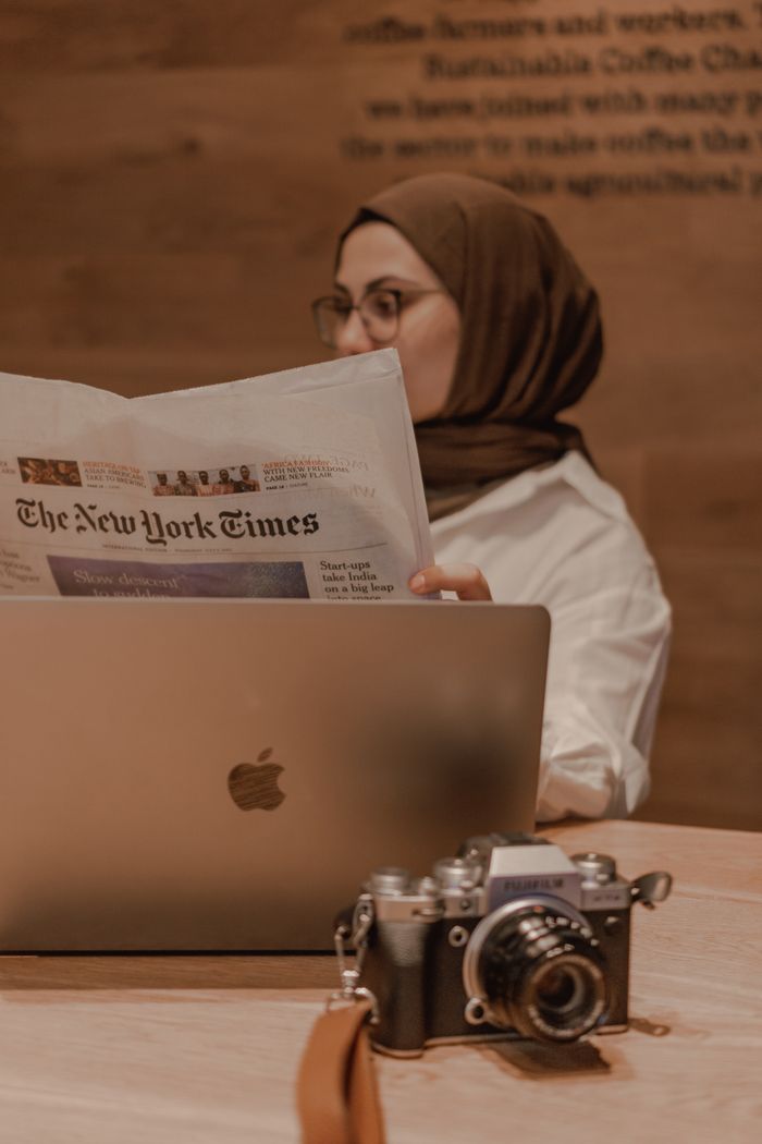 A woman in a hijab reads 'The New York Times' while sitting at a table with a MacBook and a vintage camera.